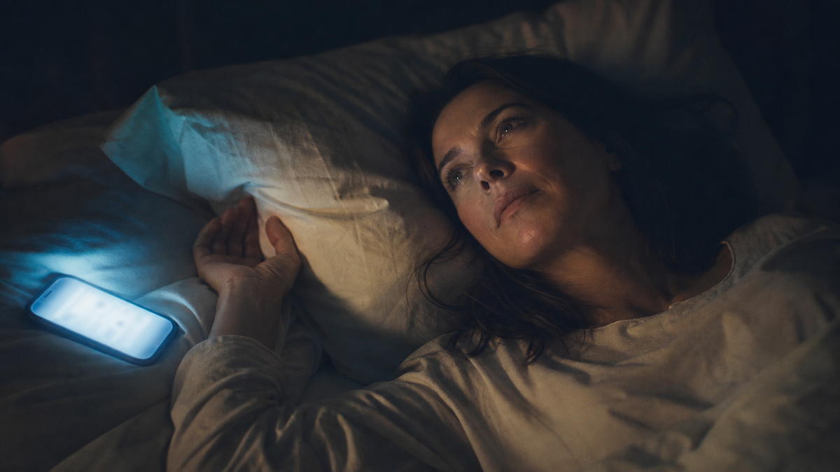 Woman in bed at 2 AM, phone on pillow face-up showing push-notification glow, eyes open staring at ceiling